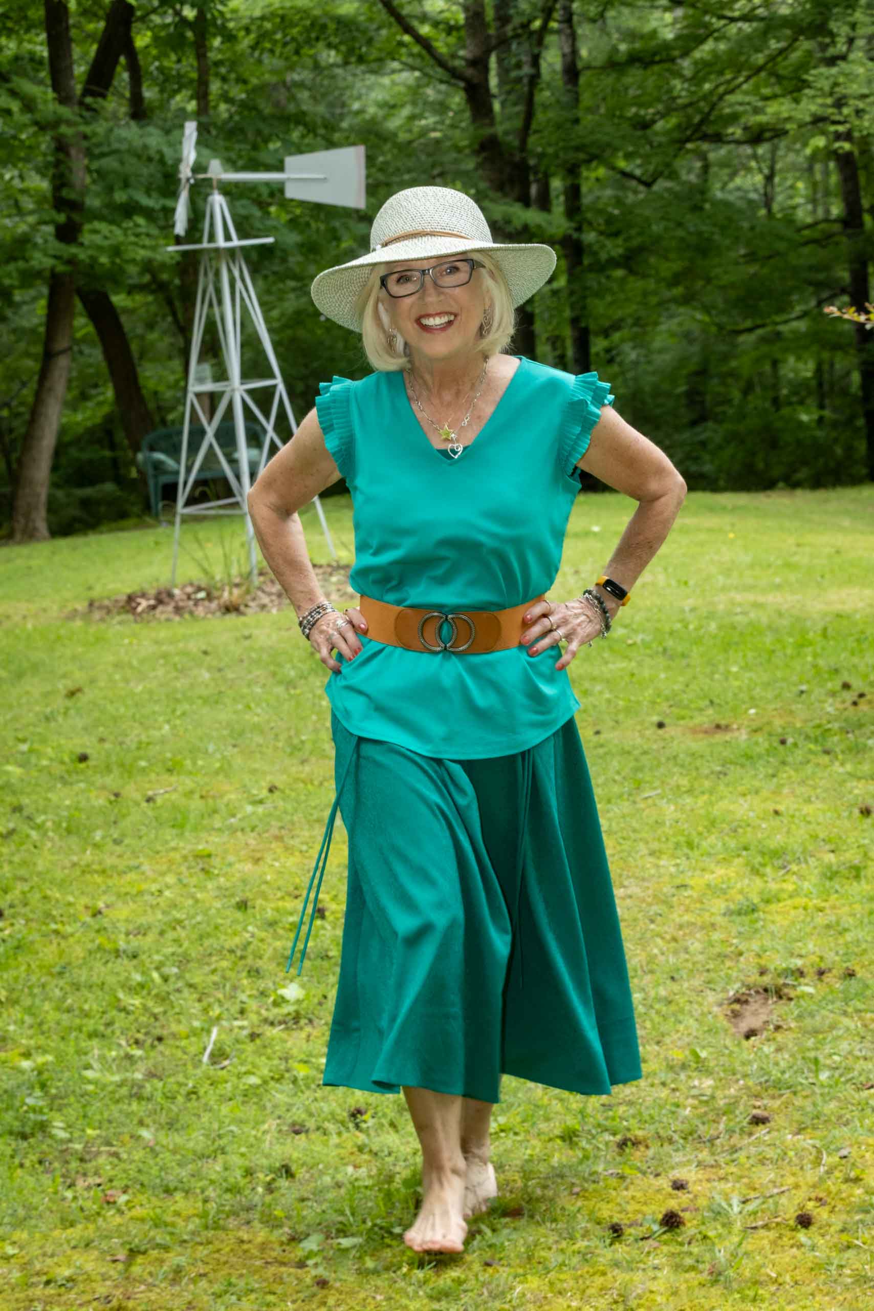 Woman in hat, green top, and green skirt walking through the grass with hands on her hips and smile on her face.